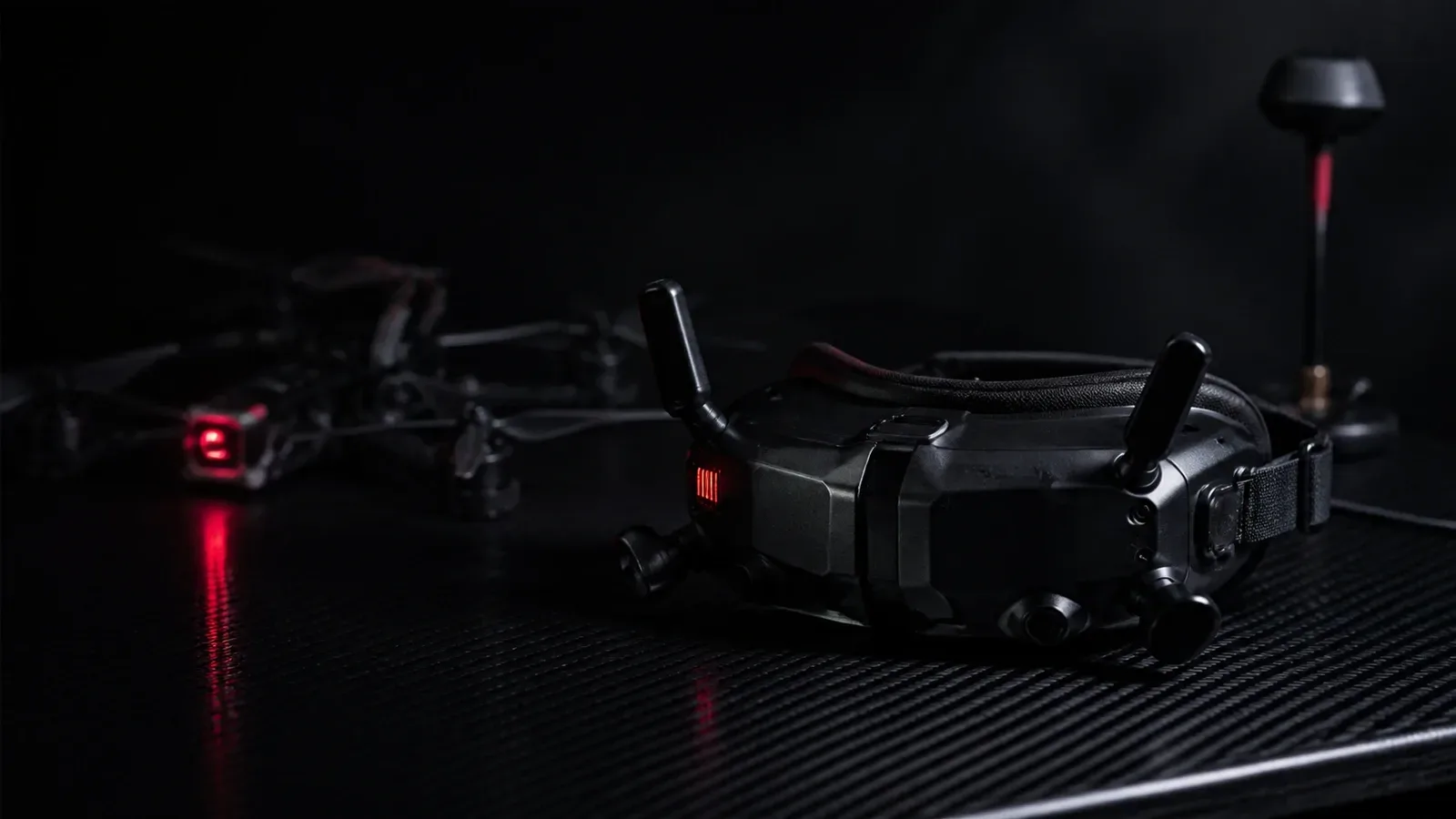 Óculos FPV de alta performance repousados sobre bancada de fibra de carbono em estúdio escuro com drone FPV ao fundo e antena de vídeo